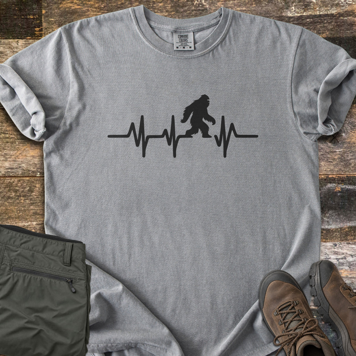 Bigfoot Heartbeat Comfort Colors T-shirt in Grey, laid flat. A black heartbeat line runs across the chest with a walking Bigfoot silhouette in the middle; there are no words. The shirt lies on weathered wood beside folded olive hiking pants and brown hiking boots.
