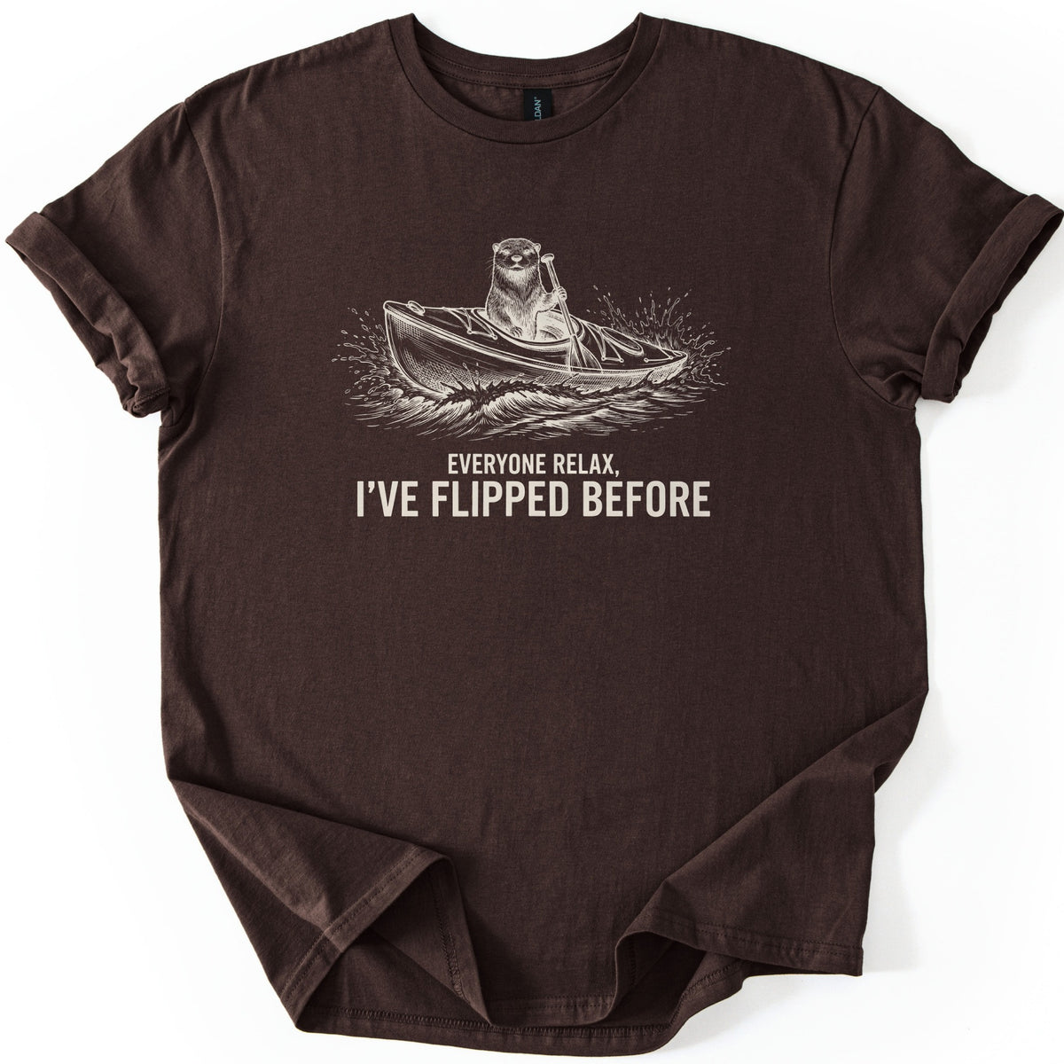 Everyone Relax I've Flipped Before Comfort Colors T-shirt in Dark Chocolate, laid flat. A cream sketch-style otter paddles a kayak through splashing waves above the words Everyone Relax, I've Flipped Before in cream. The shirt has rolled sleeves and lies on a white background.