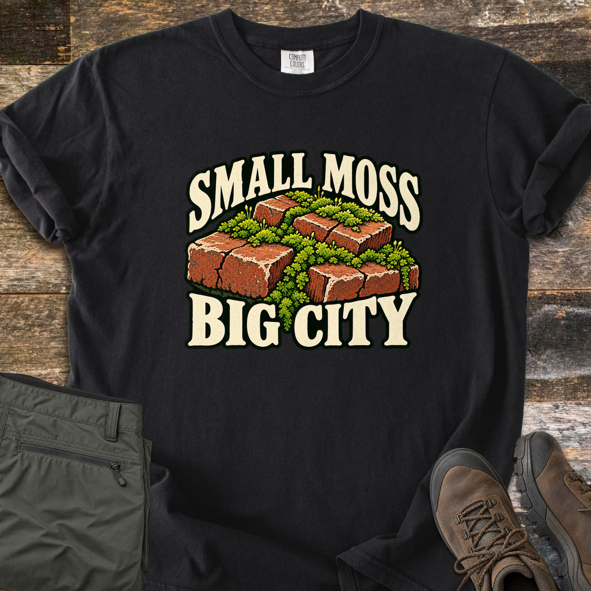 Small Moss Big City Comfort Colors T-shirt in Black, laid flat. Red bricks with bright green moss growing between and over them, with the words Small Moss Big City in cream lettering. Sleeves are rolled, on a wood floor beside folded olive pants and brown hiking boots.