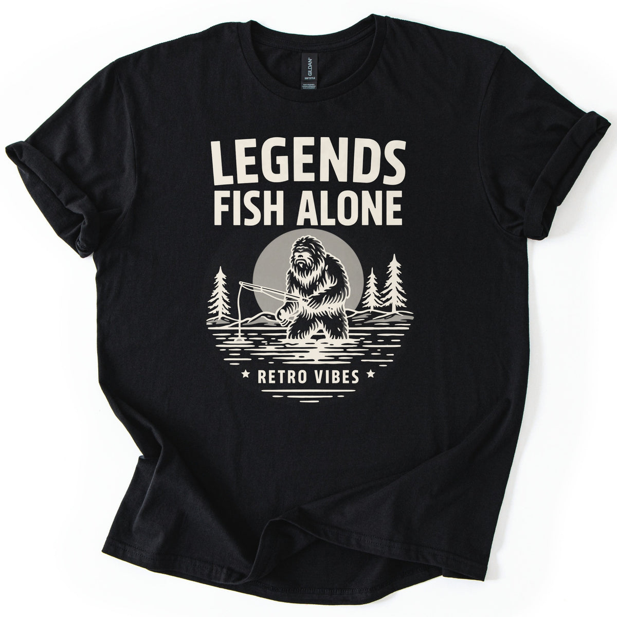 Legends Fish Alone Bigfoot Comfort Colors T-shirt in Black, laid flat. Cream print of Bigfoot fishing knee-deep in a lake before a gray moon and pine trees, under the words Legends Fish Alone and above Retro Vibes between two stars. Sleeves cuffed, on a white background.