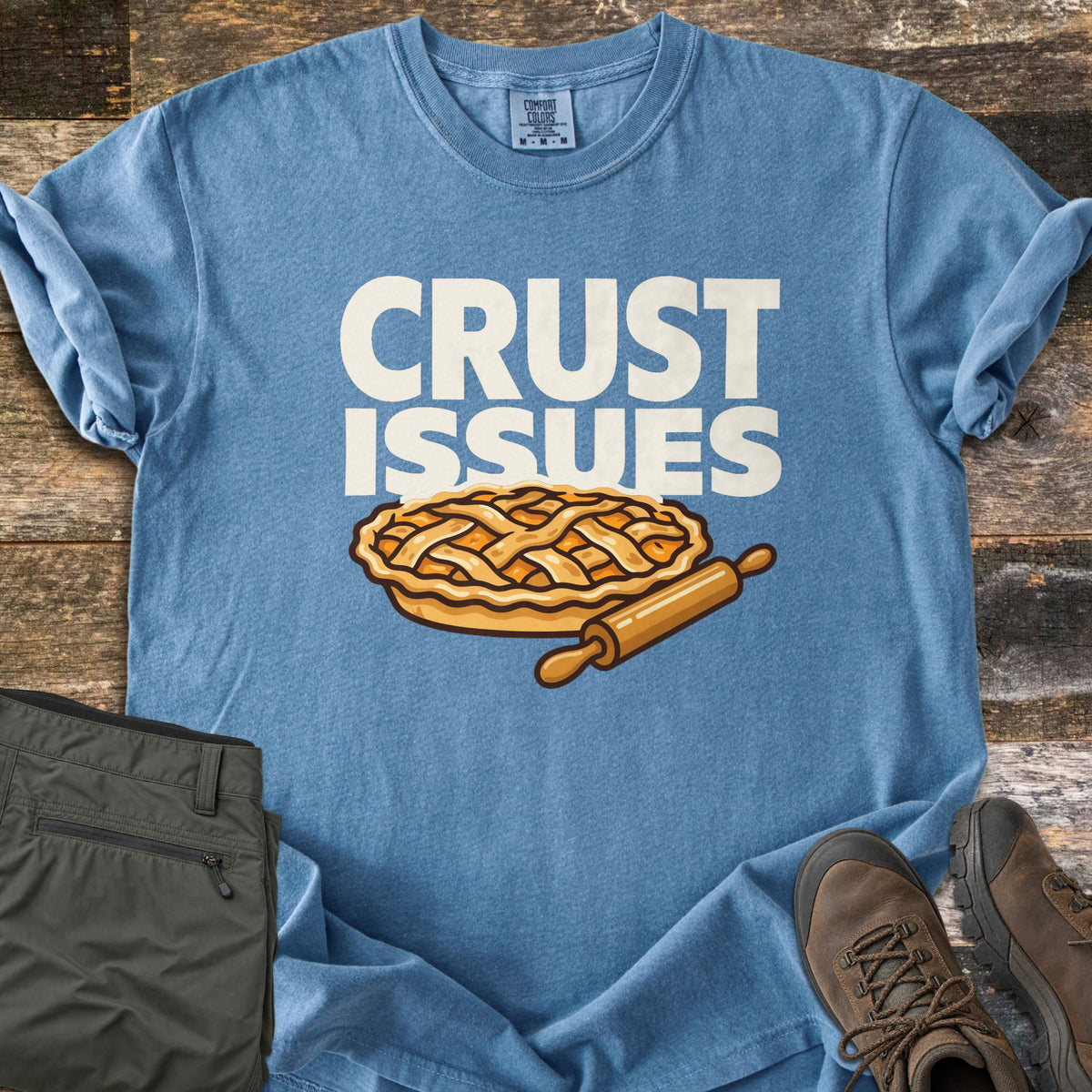 Crust Issues Comfort Colors T-shirt in Blue Jean, laid flat. The words CRUST ISSUES in off-white block letters sit above a cartoon lattice-top pie and a wooden rolling pin. Olive pants and brown hiking boots lie beside it on a rustic wood surface.