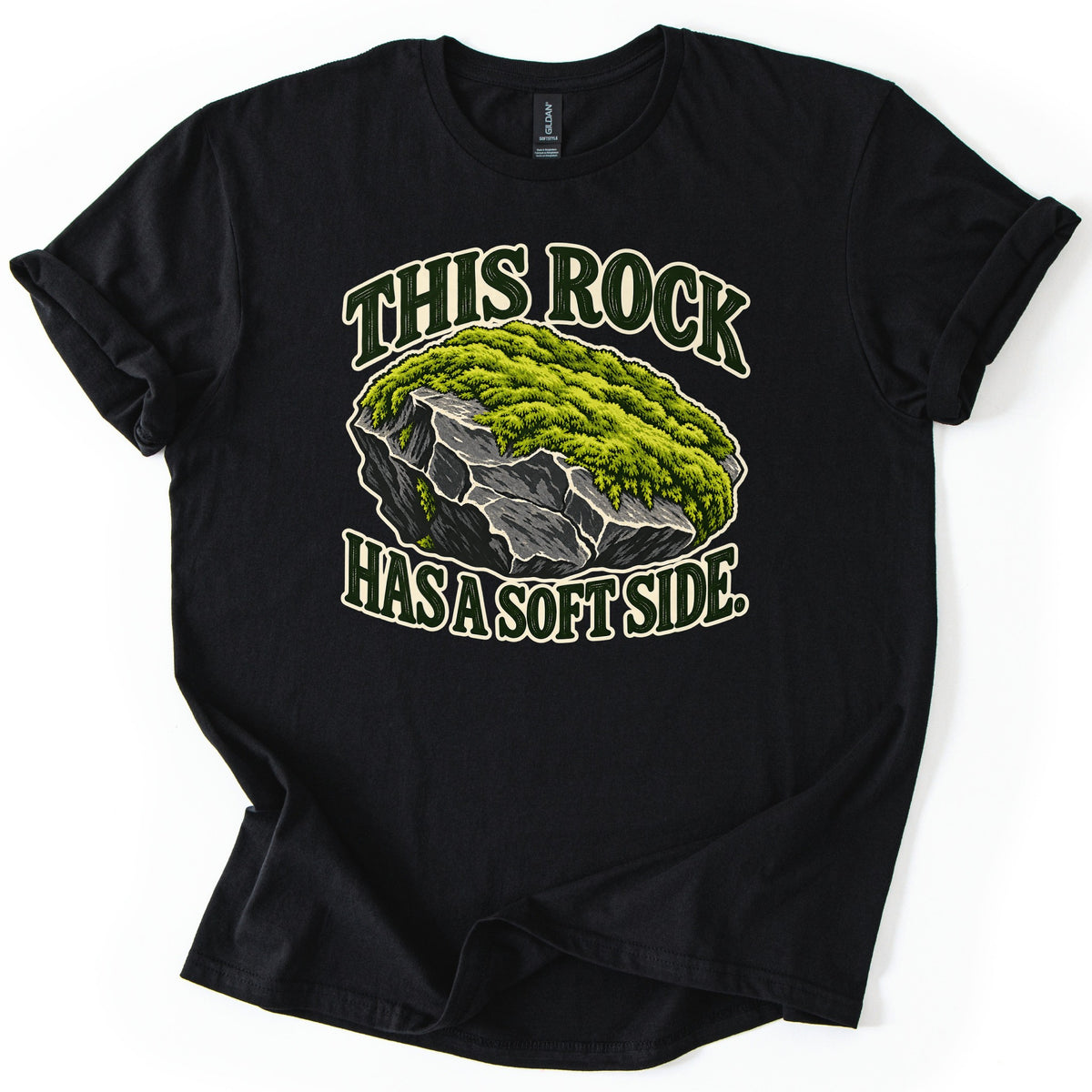 This Rock Has a Soft Side Comfort Colors T-shirt in Black, laid flat. A gray boulder topped with thick green moss sits between the words This Rock above and Has a Soft Side. below in dark green letters with a cream outline. The shirt lies on white with rolled sleeves.