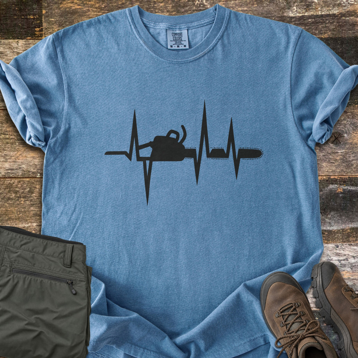 Chainsaw Heartbeat Comfort Colors T-shirt in Blue Jean, laid flat. A black heartbeat line runs across the chest with a chainsaw silhouette built into it, the bar and chain extending to the right. Folded olive cargo shorts and brown hiking boots sit on a weathered wood floor.