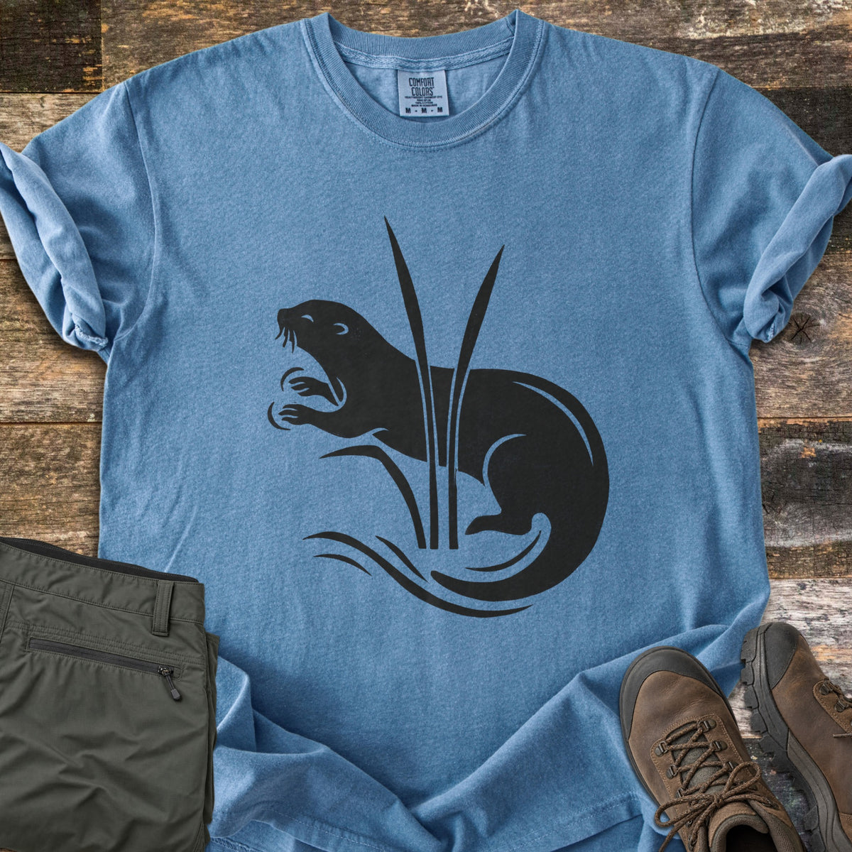 Otter In The Reeds Comfort Colors T-shirt in Blue Jean, laid flat. A black silhouette of an otter with open mouth and raised paws curls behind two tall reeds above wavy water lines, no words. Folded olive hiking pants and brown hiking boots lie on weathered wood.