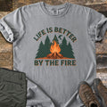 Life Is Better By The Fire Comfort Colors T-shirt in Grey, laid flat. Arched dark green text reads Life Is Better over pine trees, a crescent moon and a campfire on crossed logs, with By The Fire below. Folded olive pants and brown hiking boots sit on weathered wood.