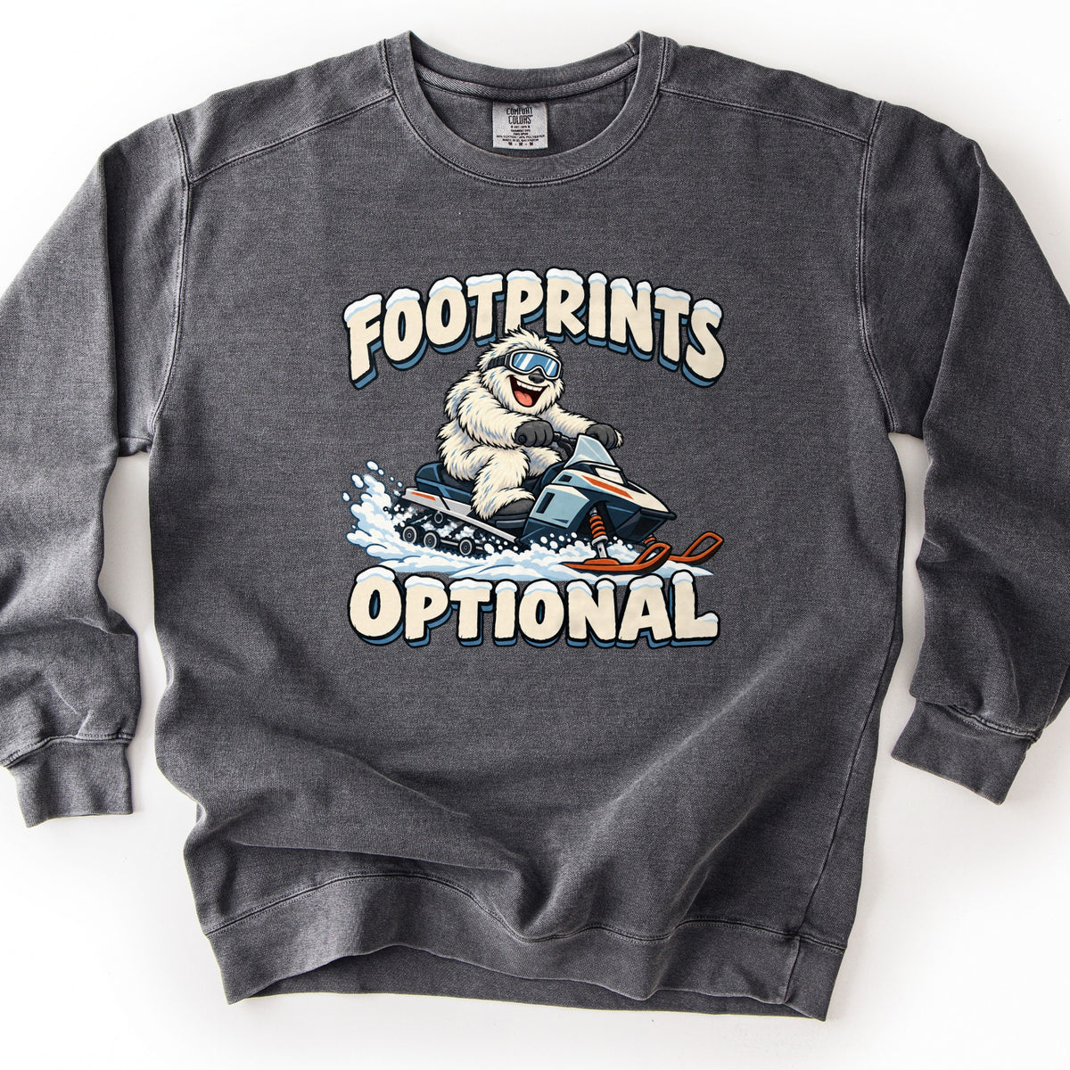 Footprints Optional Comfort Colors crewneck sweatshirt in Pepper, laid flat. A smiling white yeti in ski goggles speeds on a snowmobile through a spray of snow, between arched snow-topped cream letters reading FOOTPRINTS and OPTIONAL. Shown on a white background.