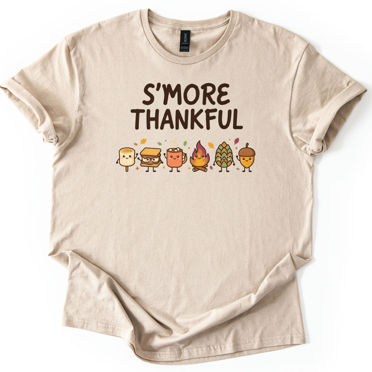Smore Thankful Comfort Colors T-shirt in Sand, laid flat. Dark brown letters read S'MORE THANKFUL above a row of smiling cartoon characters: a marshmallow, a s'more, a cocoa mug, a campfire, a pinecone and an acorn among autumn leaves. Sleeves rolled, on white.