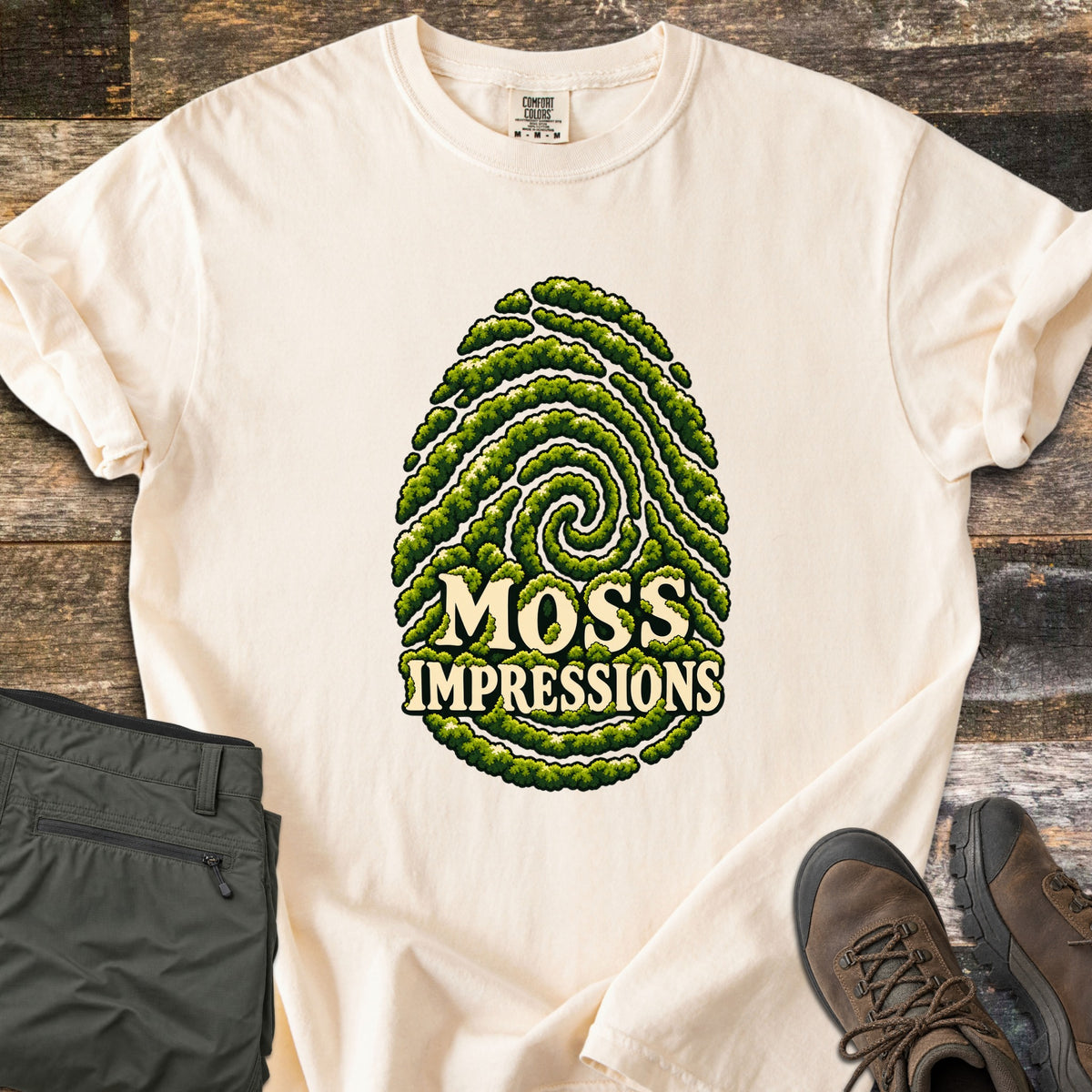 Moss Impressions Comfort Colors T-shirt in Ivory, laid flat. A fingerprint made of green moss ridges with the words Moss Impressions in cream letters. The shirt lies on weathered wood with folded olive hiking pants and brown hiking boots.