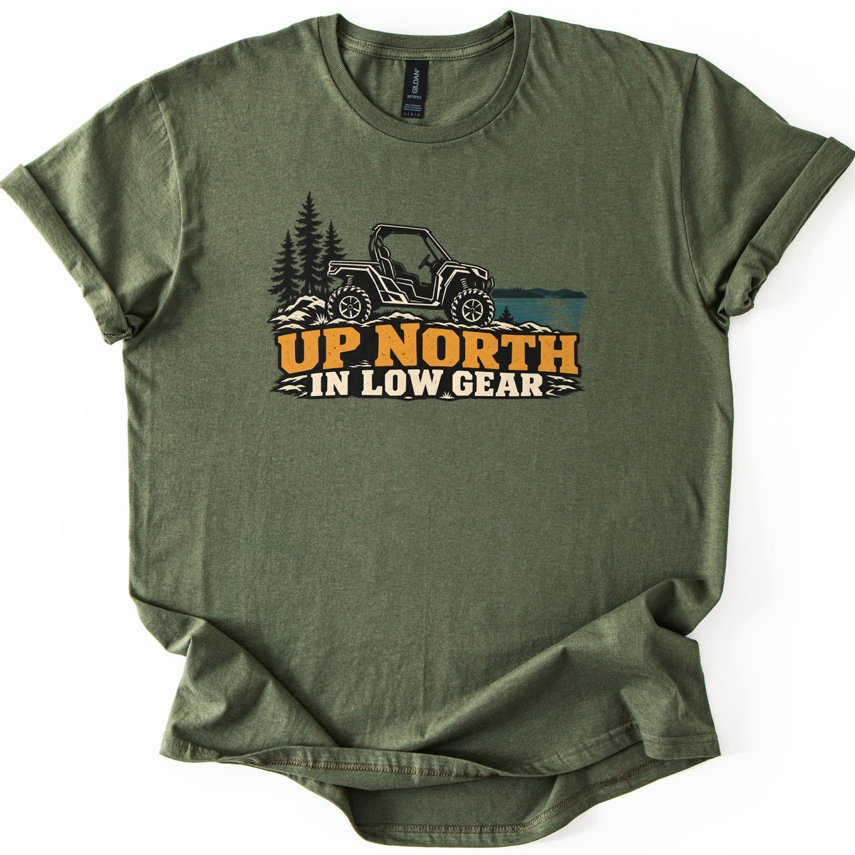 Up North in Low Gear Comfort Colors T-shirt in Military Green, laid flat. A black side-by-side UTV on rocky ground between pine trees and a blue lake sits above the words UP NORTH in orange and IN LOW GEAR in cream. The shirt lies on a white surface with rolled sleeves.