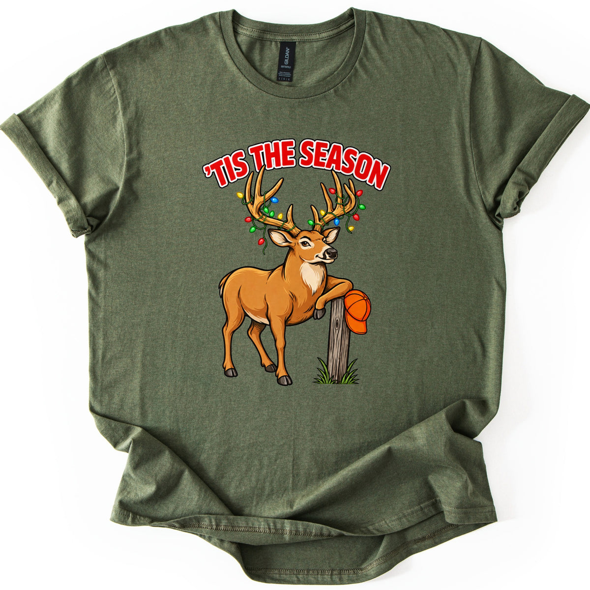 Buck Tis The Season Comfort Colors T-shirt in Military Green, laid flat. Red arched text reads 'TIS THE SEASON above a cartoon buck with colored Christmas lights strung on its antlers, leaning on a wooden post that holds an orange hunting cap. Sleeves rolled, on a white surface.