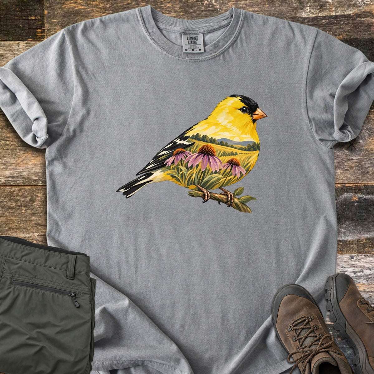 Goldfinch Meadow Comfort Colors T-shirt in Grey, laid flat. A yellow and black goldfinch on a twig, its body showing a meadow of purple coneflowers under rolling hills and a low sun. No text. Olive cargo pants and brown hiking boots lie beside it on weathered wood.