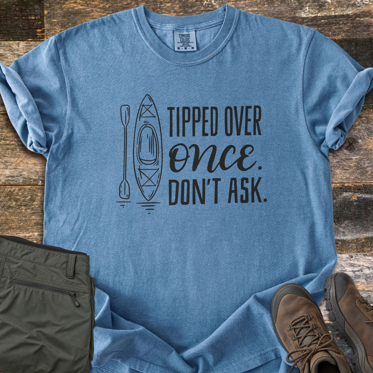 Tipped Over Once. Don't Ask. Comfort Colors T-shirt in Blue Jean, laid flat. A black line drawing of a kayak and paddle sits beside the words Tipped Over Once. Don't Ask., with Once in script. Sleeves rolled, on weathered wood with folded olive hiking pants and brown hiking boots.