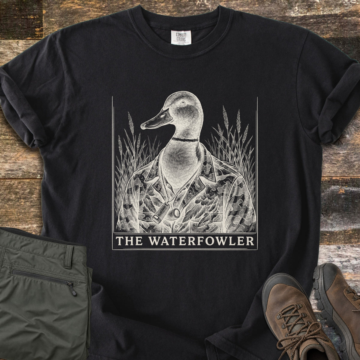 The Waterfowler Comfort Colors T-shirt in Black, laid flat. A cream engraving-style mallard in a camo jacket with a duck call on a cord stands in marsh grass above the words The Waterfowler. Folded olive pants and brown hiking boots sit beside it on weathered wood.