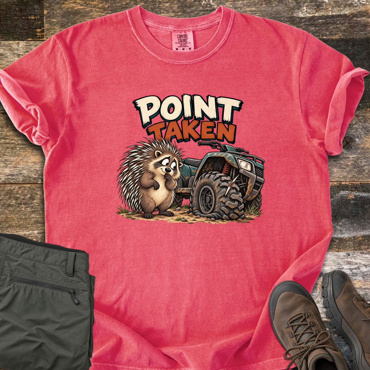 Point Taken Comfort Colors T-shirt in Watermelon, laid flat. A cartoon porcupine looks sheepishly at a muddy ATV with a flat front tire pierced by a quill, under the words Point Taken. Folded olive pants and brown hiking boots sit beside it on weathered wood.