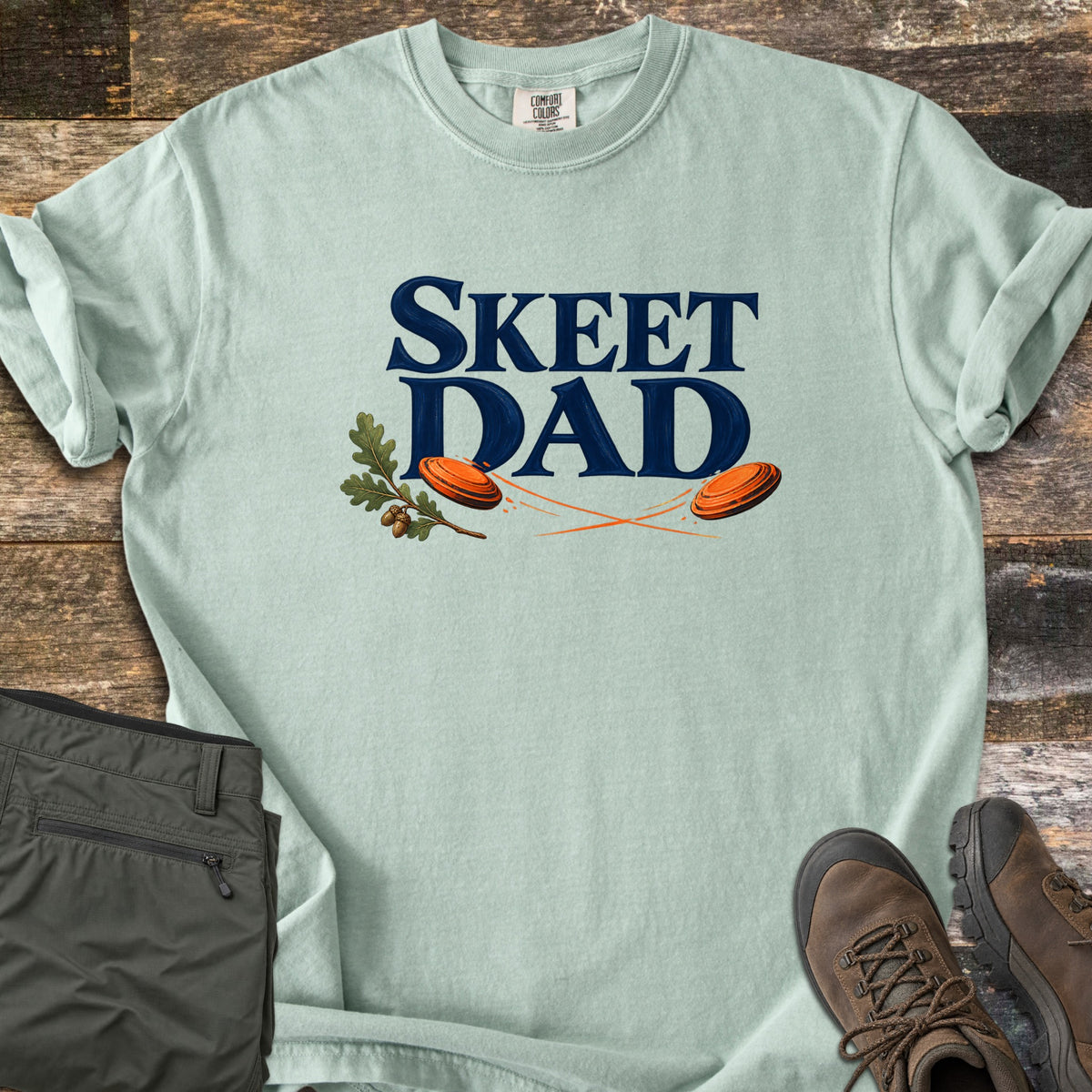 Skeet Dad Comfort Colors T-shirt in Bay, laid flat. Navy text reads Skeet Dad above two orange clay targets with motion trails and an oak sprig with acorns. Sleeves are rolled on a wood floor beside folded olive cargo pants and brown hiking boots.
