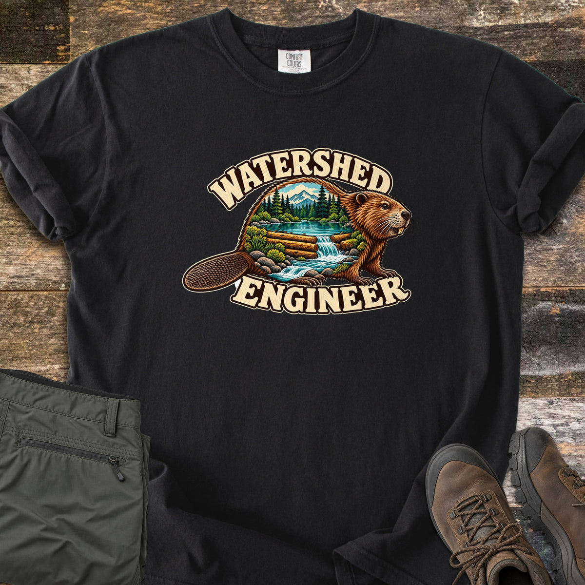 Watershed Engineer Comfort Colors T-shirt in Black, laid flat. A beaver whose body frames a forest scene with mountains, a lake, and a waterfall flowing over a log dam, between the words Watershed Engineer. Olive pants and brown hiking boots lie on a wood surface.