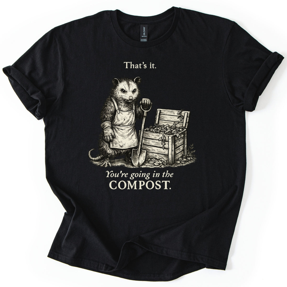 Going In The Compost Possum Comfort Colors T-shirt in Black, laid flat. A scowling possum in an apron and work glove leans on a shovel beside an open wooden compost bin, between the words That's it. and You're going in the COMPOST. Cream print, shirt on white with cuffed sleeves.