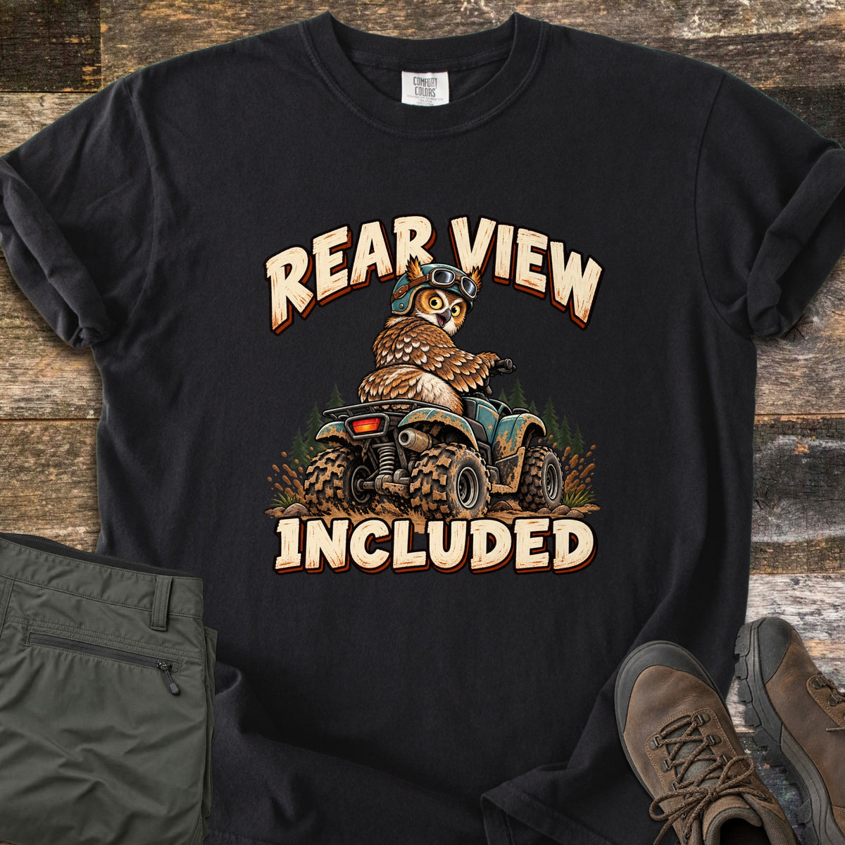 Rear View Included Comfort Colors T-shirt in Black, laid flat. A cartoon owl in a helmet and goggles rides a muddy ATV past pine trees, looking back over its shoulder, between the words Rear View Included. Olive cargo pants and brown hiking boots lie on a wood floor.