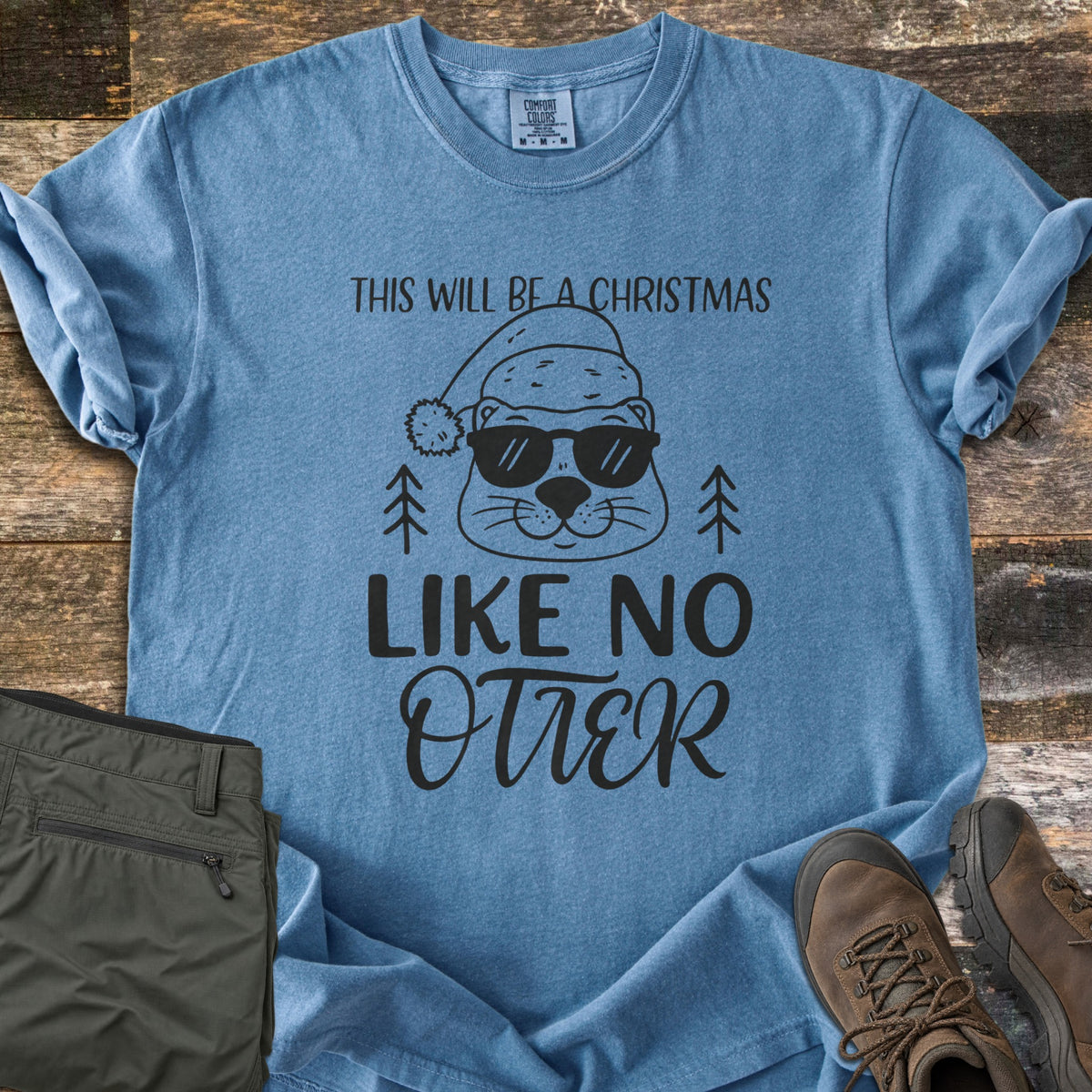 Christmas Like No Otter Comfort Colors T-shirt in Blue Jean, laid flat. A cartoon otter in a Santa hat and sunglasses sits between two pine trees, with This Will Be a Christmas above and Like No Otter below, in black. Olive cargo pants and brown hiking boots lie on weathered wood.
