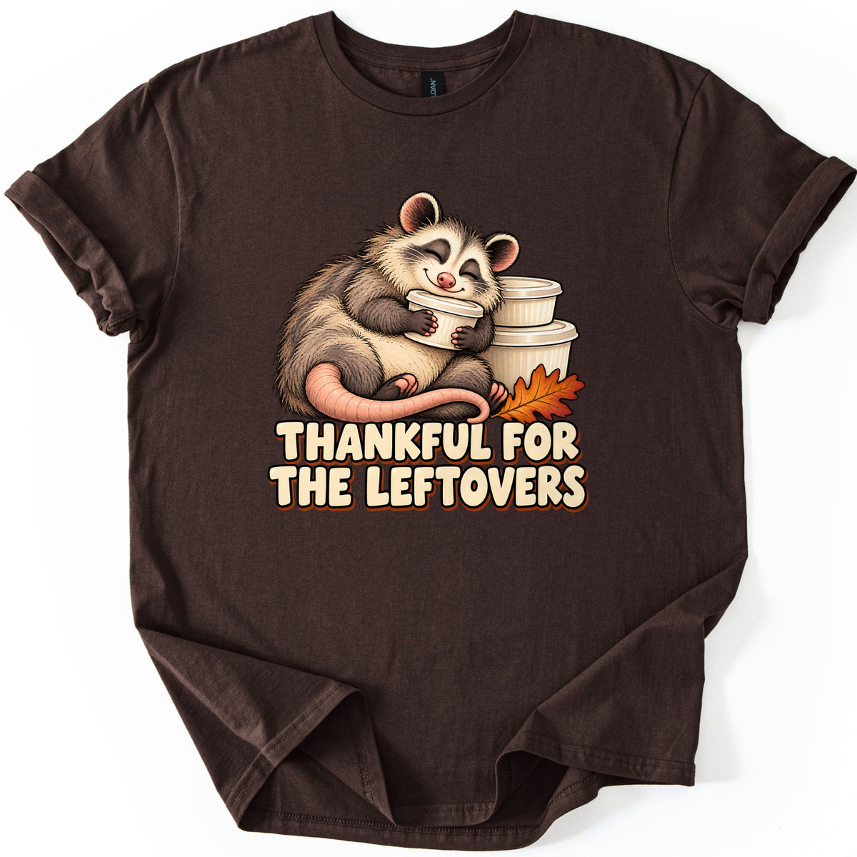 Thankful For The Leftovers Comfort Colors T-shirt in Dark Chocolate, laid flat. A sleepy, smiling opossum hugs a white leftovers container beside stacked containers and an orange oak leaf, above cream bubble letters reading Thankful For The Leftovers. Sleeves cuffed on white.