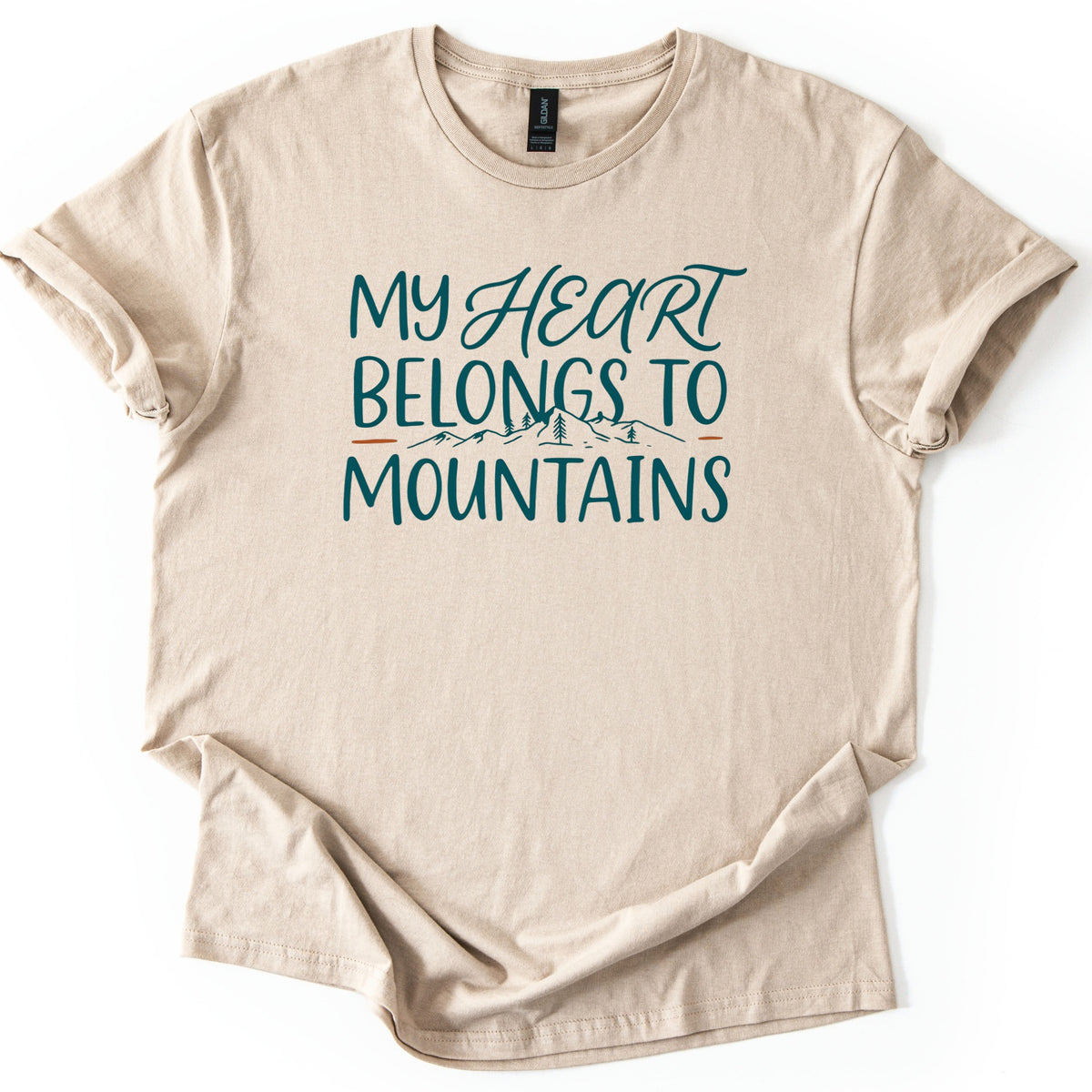 My Heart Belongs to Mountains Comfort Colors T-shirt in Sand, laid flat. Teal lettering reads My Heart Belongs to Mountains, with Heart in script above a small line drawing of mountains and pine trees between orange dashes. The shirt has cuffed sleeves and lies on a white background.