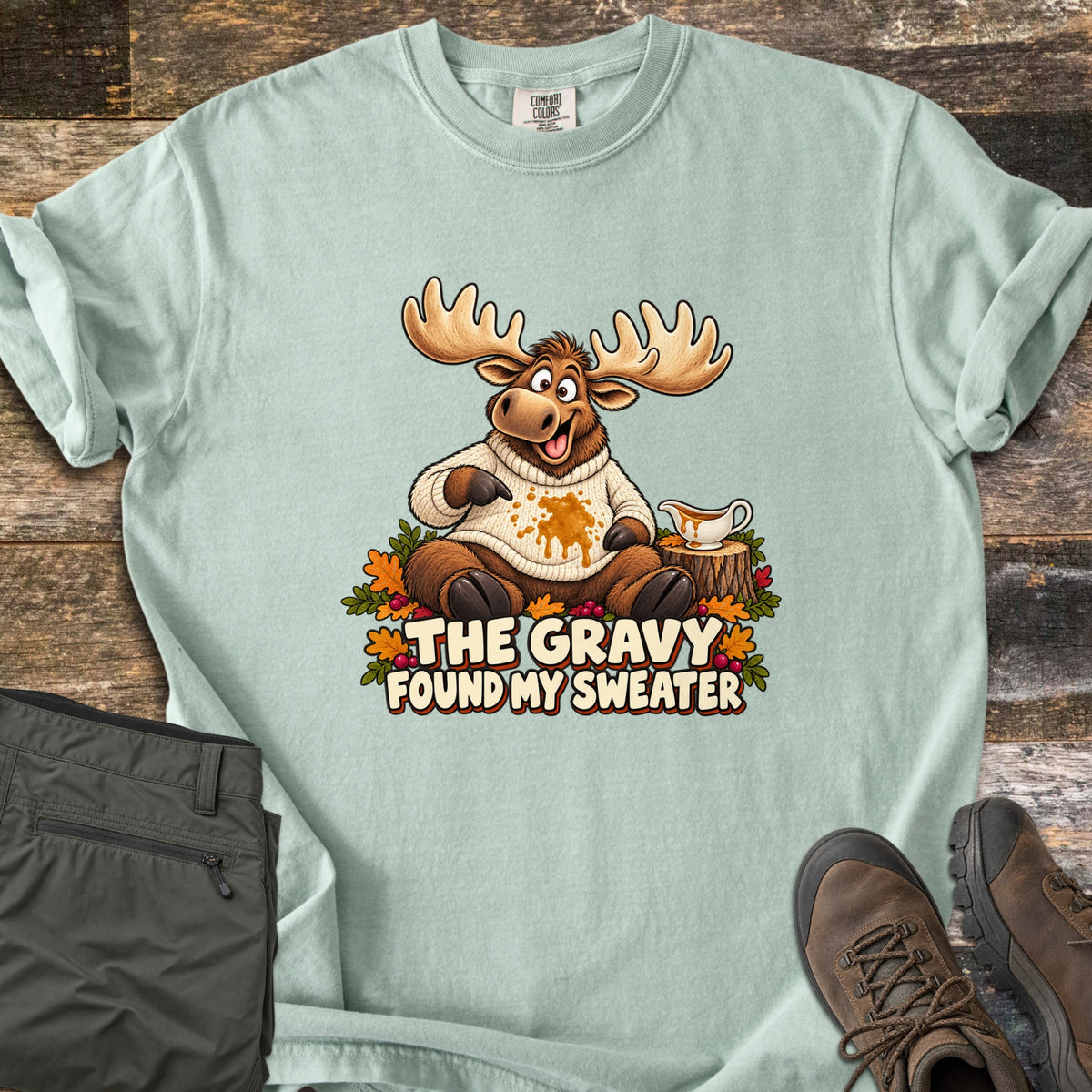 The Gravy Found My Sweater Comfort Colors T-shirt in Bay, laid flat. A cartoon moose in a cream knit sweater points at a gravy stain beside a gravy boat on a stump, above the words The Gravy Found My Sweater. Olive pants and brown boots sit on a wood floor.