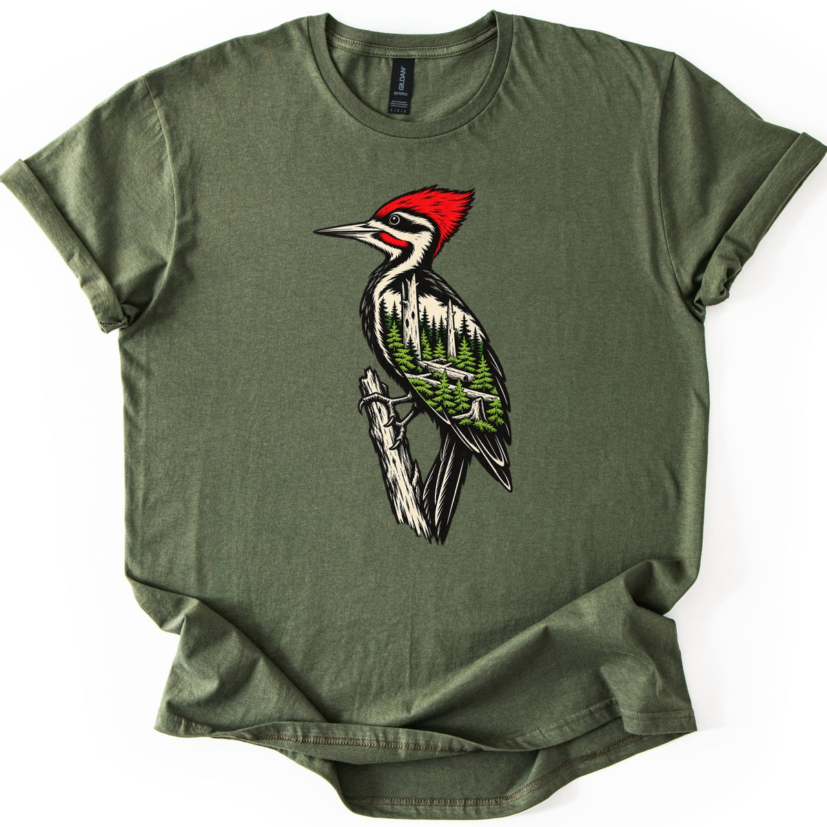Woodpecker Forest Comfort Colors T-shirt in Military Green, laid flat. A red-crested woodpecker perches on a broken branch, its black and cream body filled with a green pine forest scene. No words are printed. The sleeves are rolled and the hem is folded on a white background.