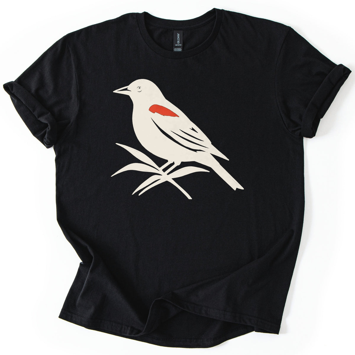 Red Winged Blackbird Comfort Colors T-shirt in Black, laid flat. A cream silhouette of a red-winged blackbird with a red shoulder patch, perched on slender leaves, with no words. The shirt has rolled sleeves and lies on a white background.