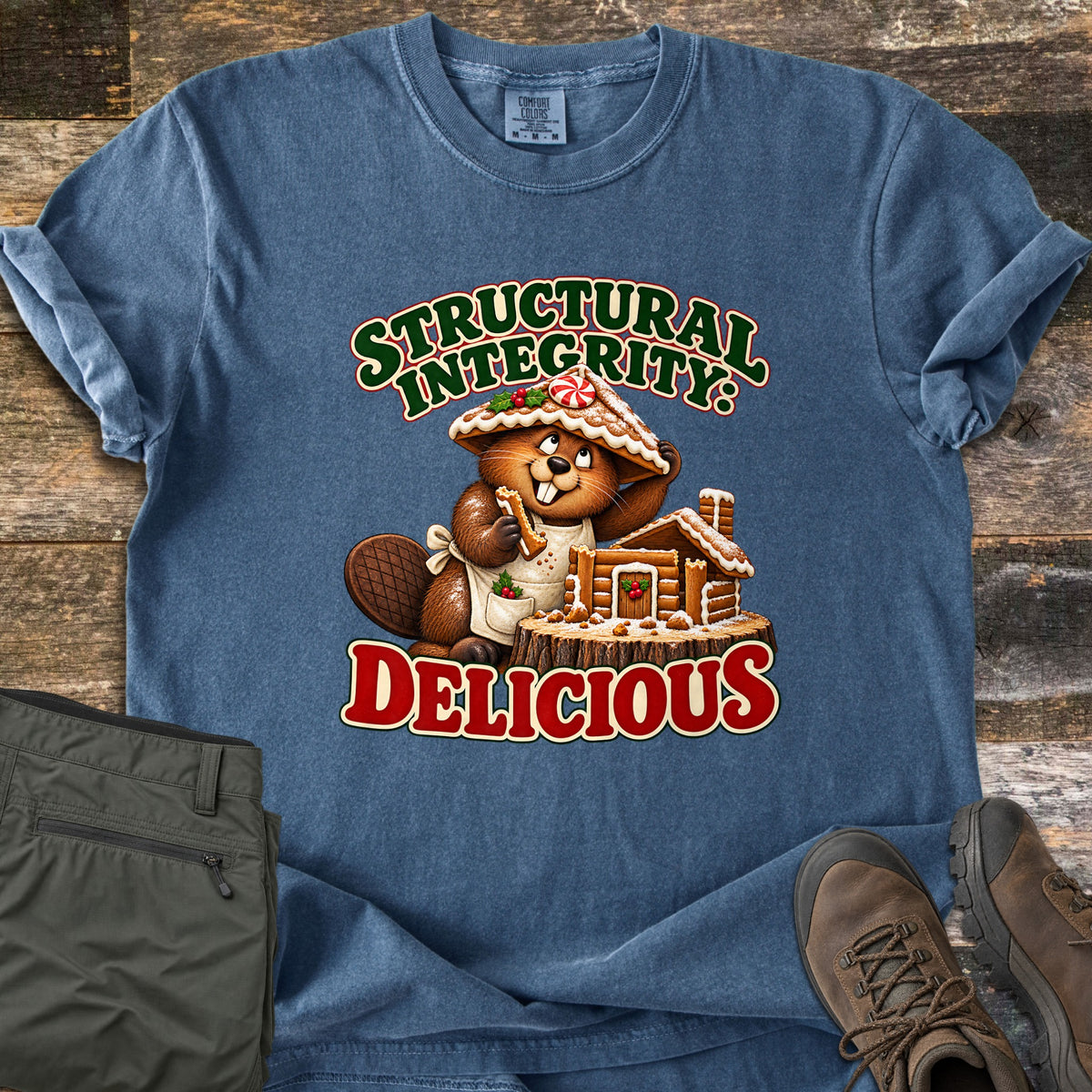 Structural Integrity: Delicious Comfort Colors T-shirt in Denim, laid flat. A cartoon beaver in an apron, wearing a gingerbread roof as a hat, nibbles a cookie beside a gingerbread cabin on a stump, between the words Structural Integrity: Delicious. Olive pants and brown boots lie on wood planks.