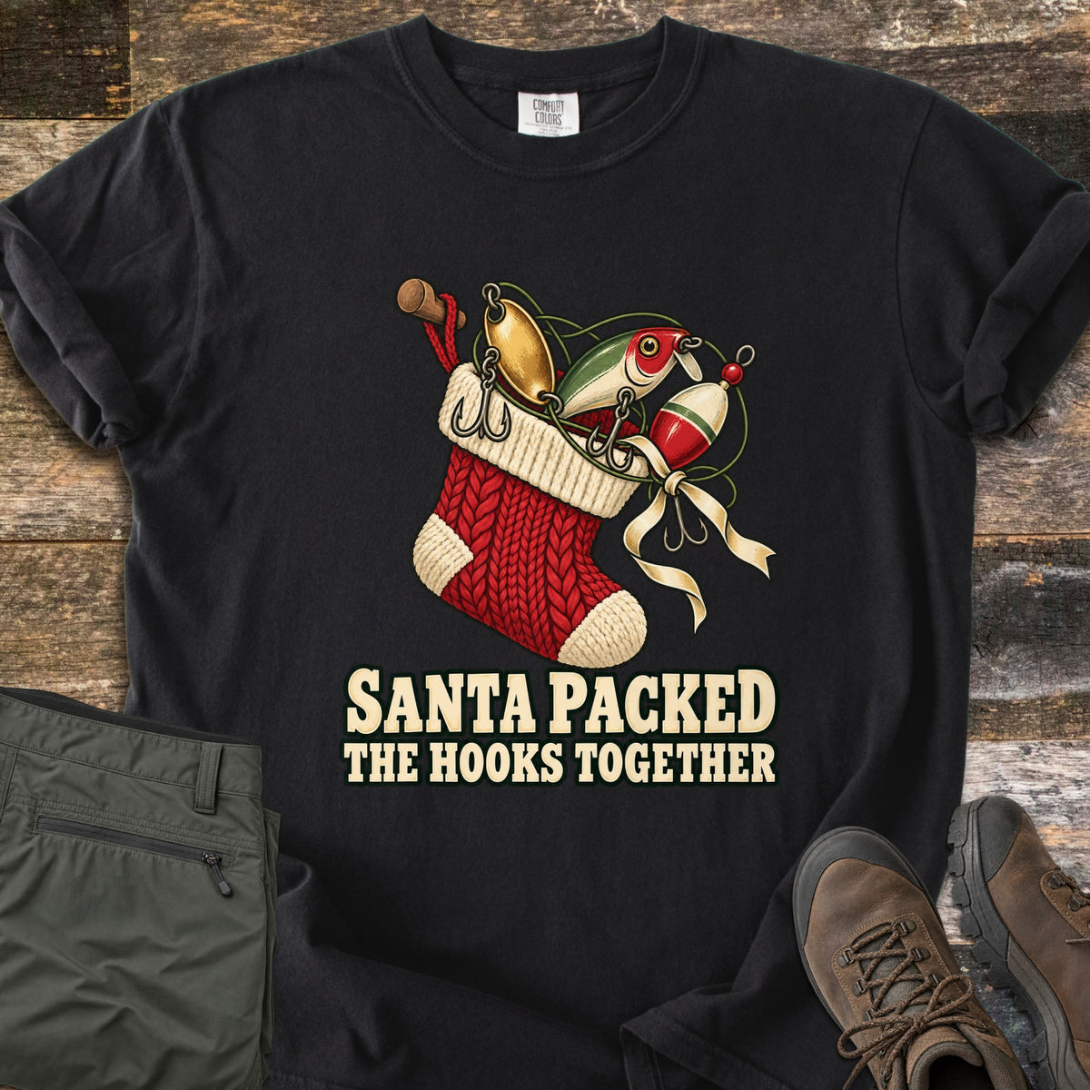 Santa Packed The Hooks Together Comfort Colors T-shirt in Black, laid flat. A red knit Christmas stocking holds fishing lures, a bobber and a rod handle tied with a cream ribbon above the words Santa Packed The Hooks Together. Olive pants and brown boots sit on a wood floor.