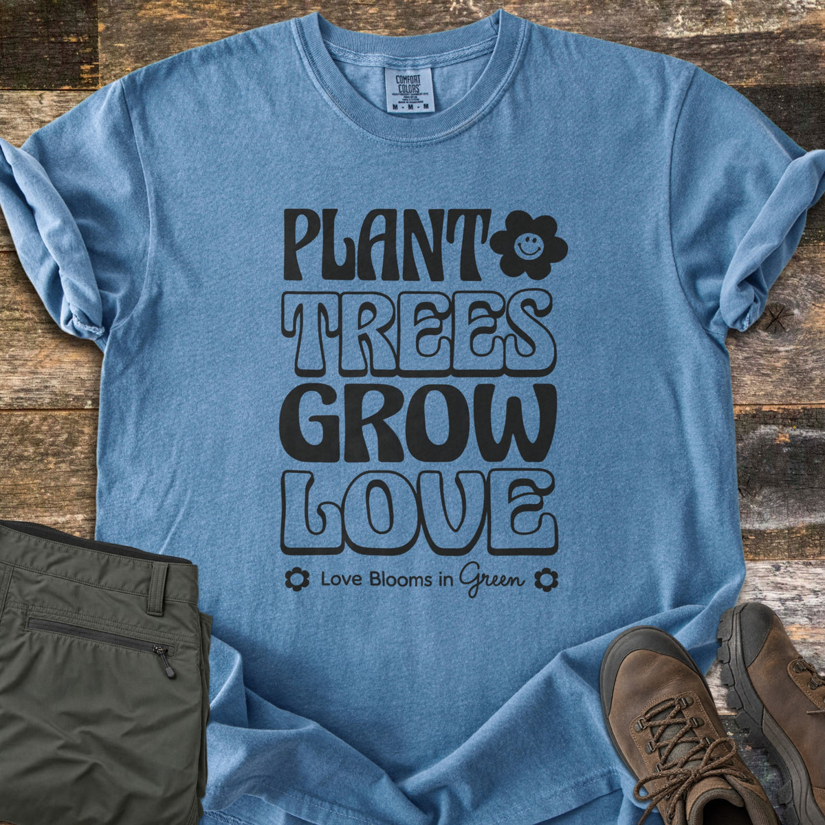 Plant Trees Grow Love Comfort Colors T-shirt in Blue Jean, laid flat. Black retro bubble letters read Plant Trees Grow Love beside a smiling daisy, above Love Blooms in Green. Sleeves cuffed on weathered wood with folded olive hiking pants and brown hiking boots.