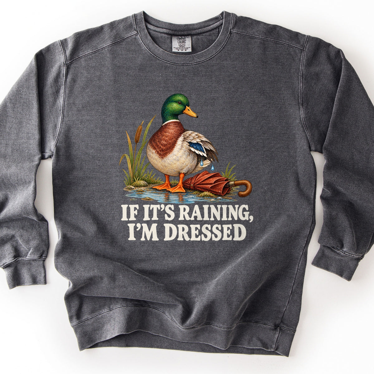 If It's Raining, I'm Dressed Comfort Colors crewneck sweatshirt in Pepper, laid flat. A mallard duck with water dripping from its wing stands in a puddle by cattails beside a closed red umbrella, above the words If It's Raining, I'm Dressed in cream. Shown on a white background.