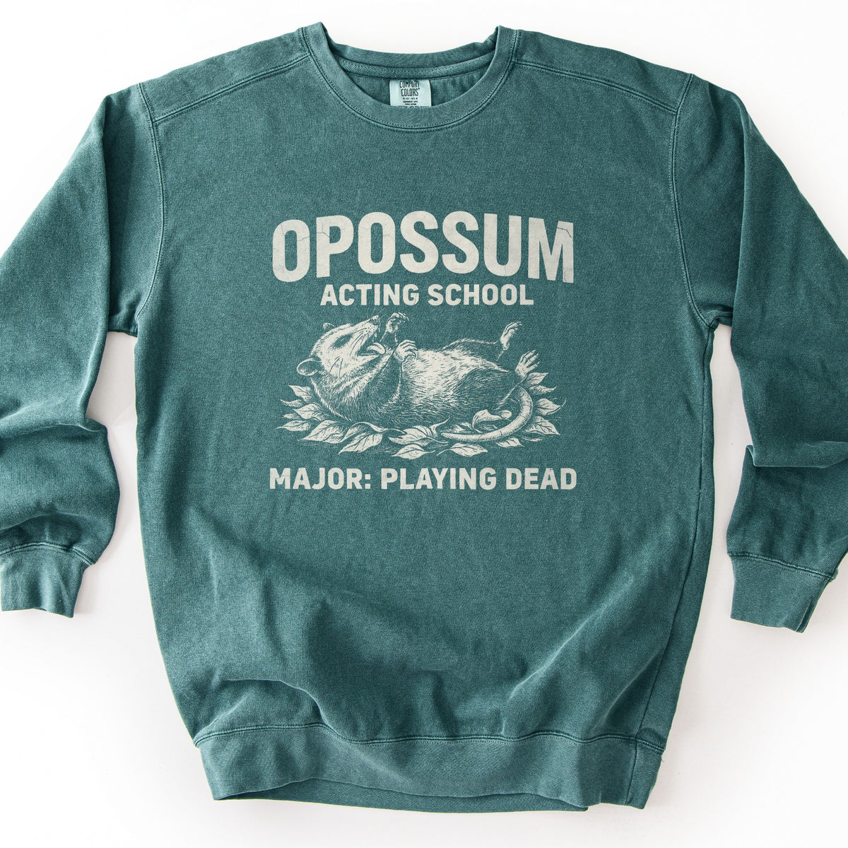 Opossum Acting School Comfort Colors crewneck sweatshirt in Blue Spruce, laid flat. Distressed off-white print of the words Opossum Acting School above an opossum lying on its back in leaves with its mouth open, and Major: Playing Dead below. Shown on a plain white background.