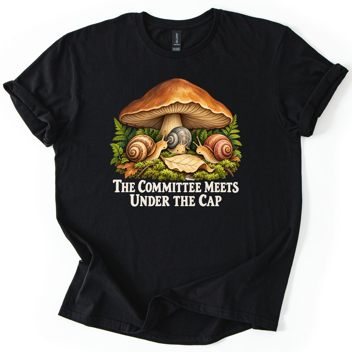 The Committee Meets Under the Cap Comfort Colors T-shirt in Black, laid flat. A large brown mushroom shelters three snails on moss among ferns and a fallen leaf, above cream words The Committee Meets Under the Cap. The shirt lies on a white background with rolled sleeves.