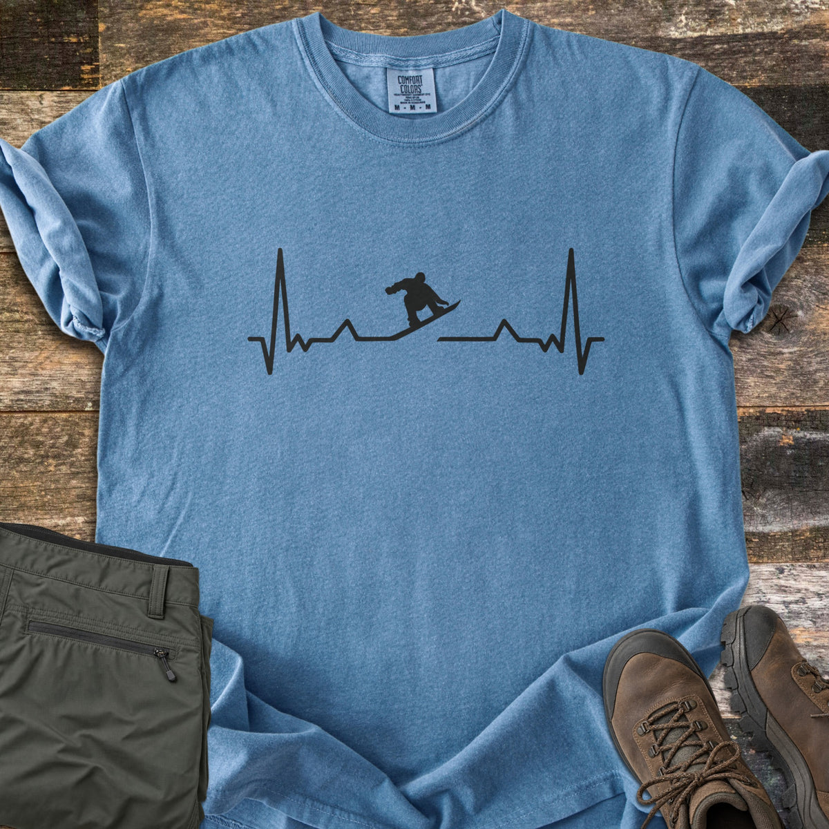 Snowboarder Heartbeat Comfort Colors T-shirt in Blue Jean, laid flat. A black heartbeat line runs across the chest, with a snowboarder silhouette riding the line in the middle. Sleeves are rolled, on a rustic wood floor beside folded olive pants and brown hiking boots.