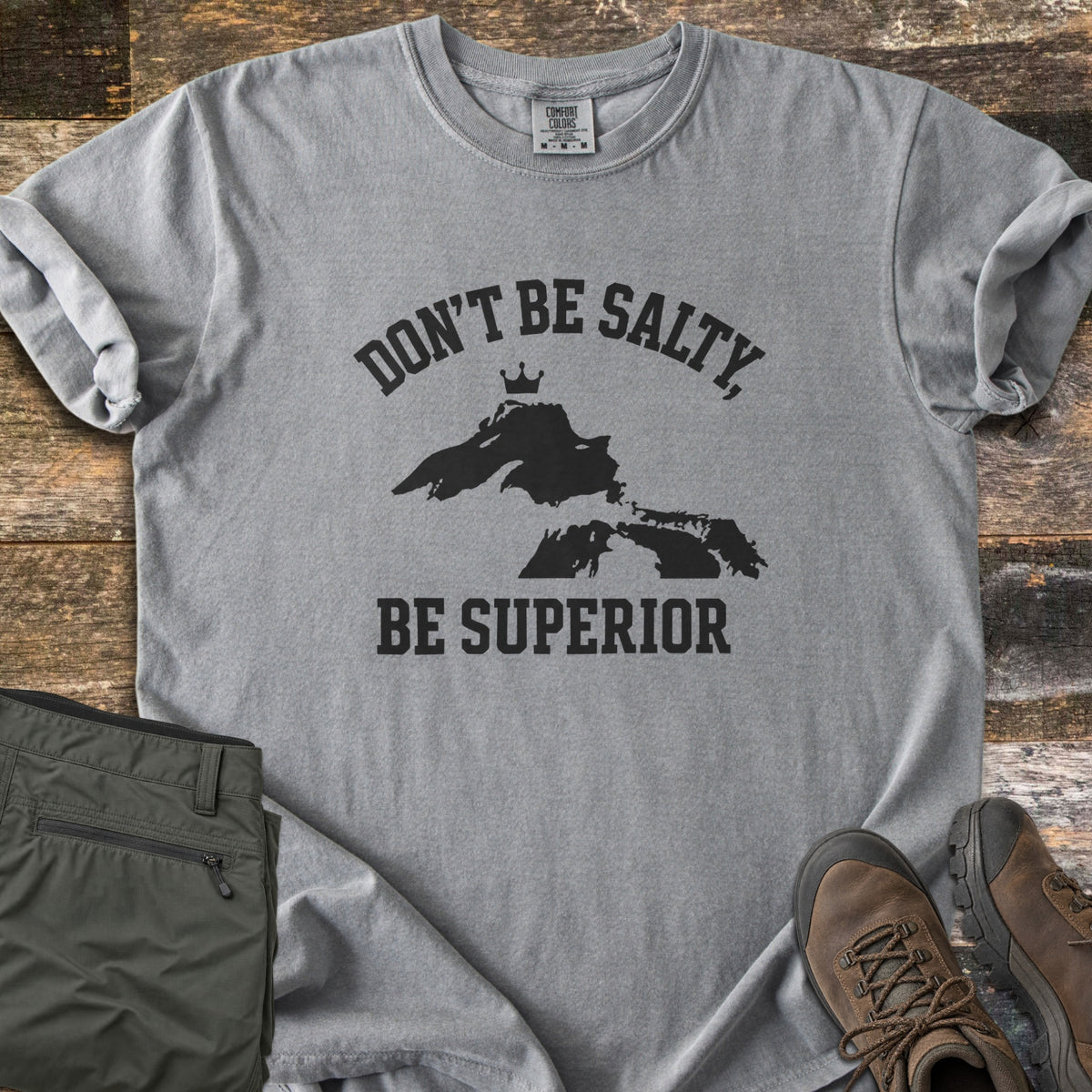 Be Superior Comfort Colors T-shirt in Grey, laid flat. Black text reads Don't Be Salty, arched over a Lake Superior silhouette topped with a small crown, with Be Superior below. Sleeves rolled, on weathered wood with olive hiking pants and brown hiking boots.