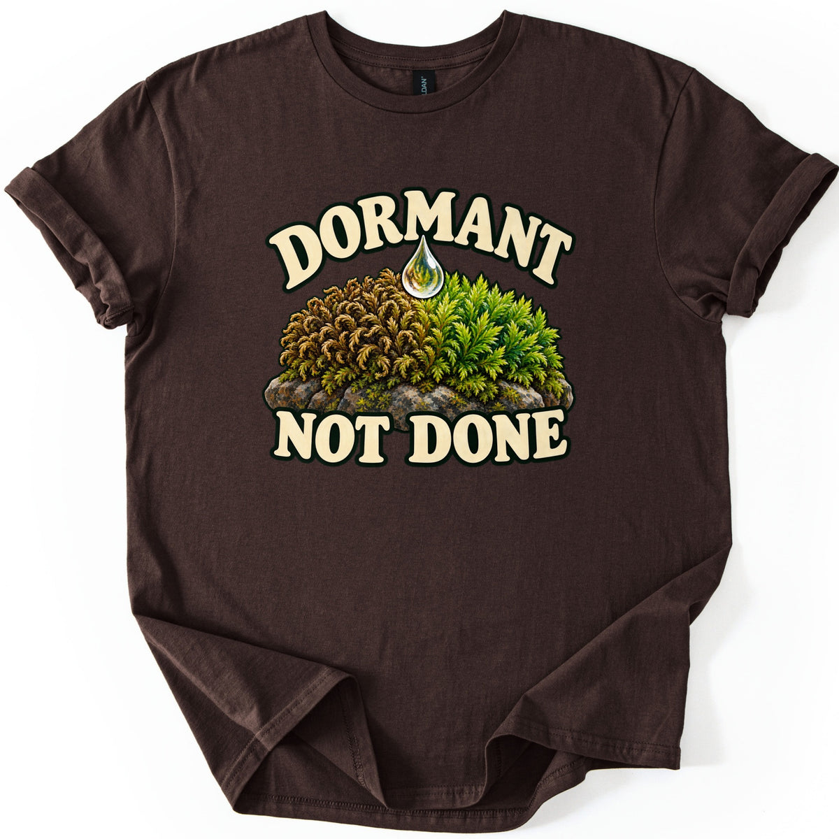 Dormant Not Done Comfort Colors T-shirt in Dark Chocolate, laid flat. The words Dormant and Not Done in cream outlined letters frame a mossy rock that turns from dry brown to fresh green beneath a falling water drop. Sleeves cuffed on a white background.