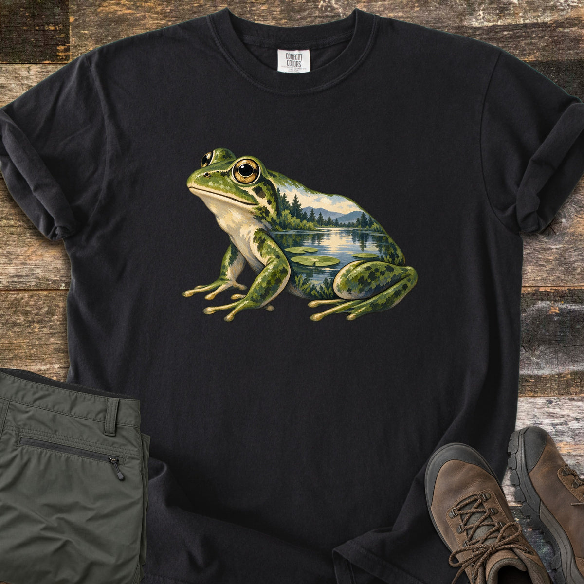 Lily Pond Frog Comfort Colors T-shirt in Black, laid flat. A painted green frog sits in profile, its body filled with a lake scene of pine trees, mountains, and lily pads. Folded olive pants and brown hiking boots lie beside it on weathered wood boards.