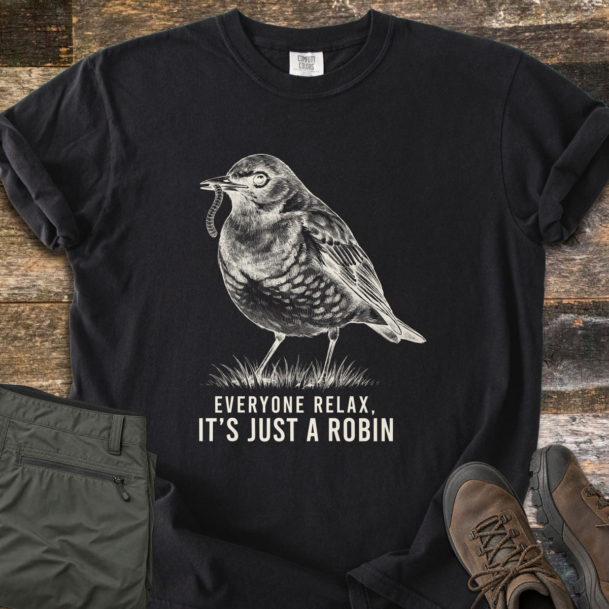 Everyone Relax It's Just A Robin Comfort Colors T-shirt in Black, laid flat. A sketched robin with a worm in its beak stands on grass above the words Everyone Relax, It's Just A Robin, in cream ink. Olive cargo pants and brown hiking boots lie beside it on weathered wood.
