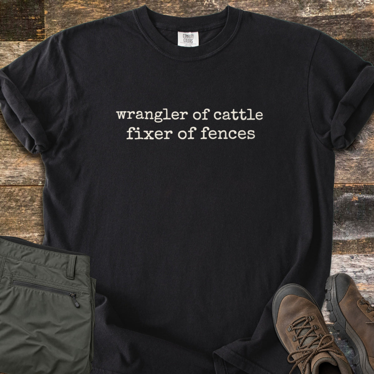 Fixer of Fences Comfort Colors T-shirt in Black, laid flat. Off-white typewriter-style text across the chest reads wrangler of cattle, fixer of fences. The shirt has rolled sleeves and lies on weathered wood planks beside folded olive green pants and brown hiking boots.