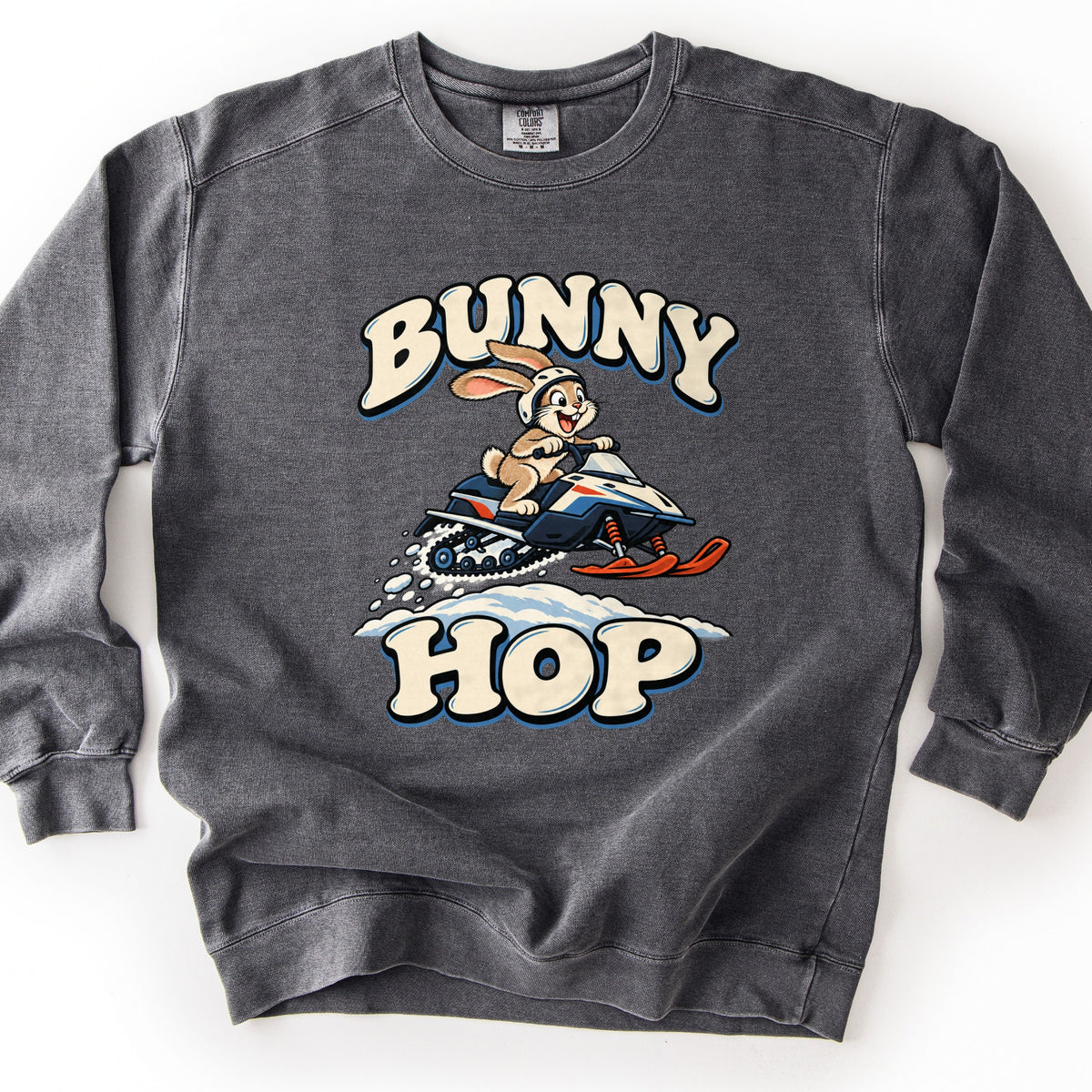 Bunny Hop Comfort Colors crewneck sweatshirt in Pepper, laid flat. A grinning cartoon bunny in a white helmet rides a navy, white and orange snowmobile over a snowbank between the words Bunny Hop in cream bubble letters. The sweatshirt lies on a white surface.