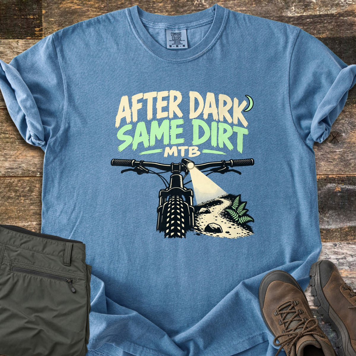 After Dark Same Dirt Comfort Colors T-shirt in Blue Jean, laid flat. Printed words After Dark Same Dirt MTB with a crescent moon above a mountain bike's handlebars, its headlight beaming onto a dirt trail with a fern. Olive pants and brown hiking boots lie on dark wood planks.