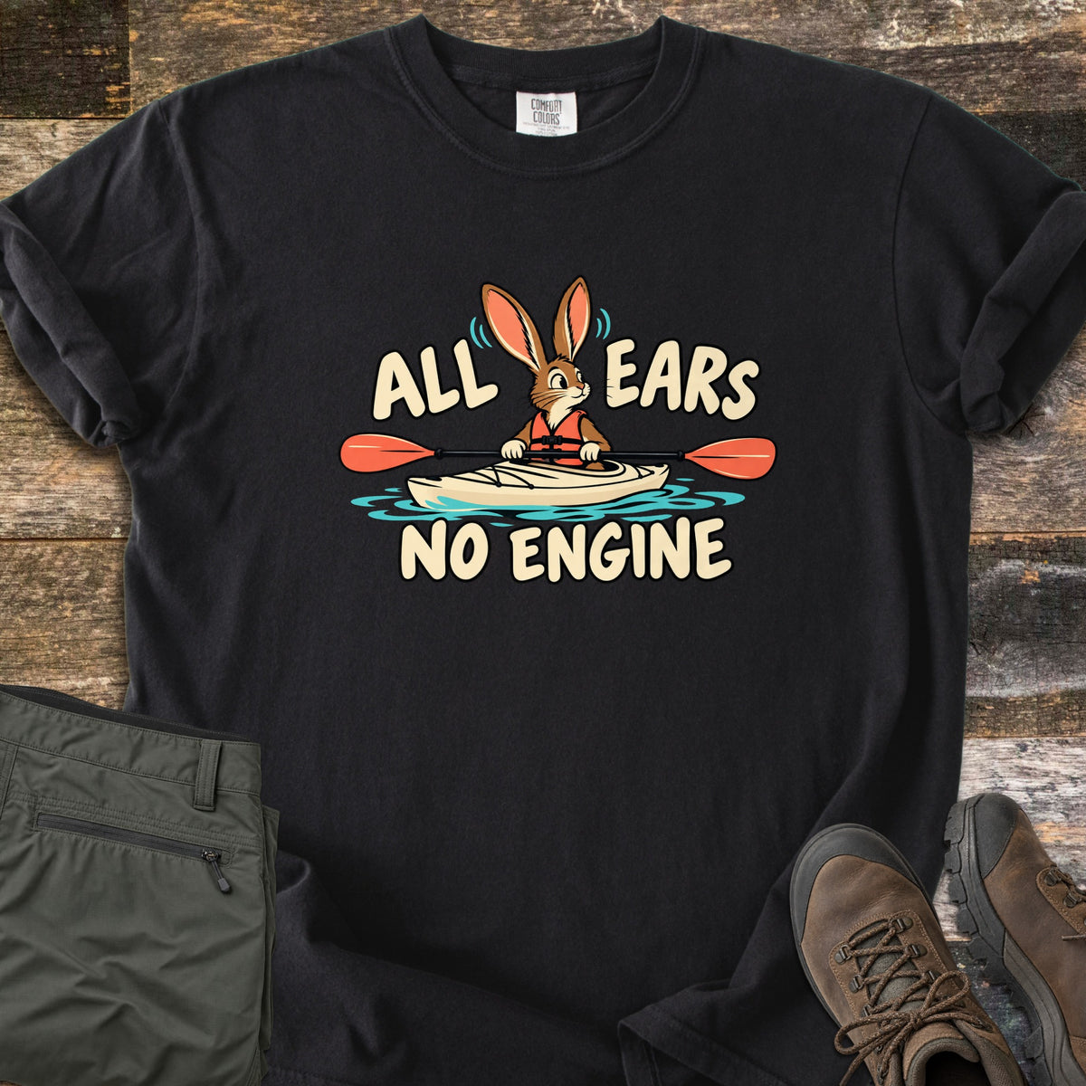 All Ears No Engine Comfort Colors T-shirt in Black, laid flat. A cartoon rabbit in an orange life vest paddles a cream kayak with an orange-bladed paddle on blue water, between the words All Ears No Engine. Olive cargo pants and brown hiking boots sit on a wood plank floor.