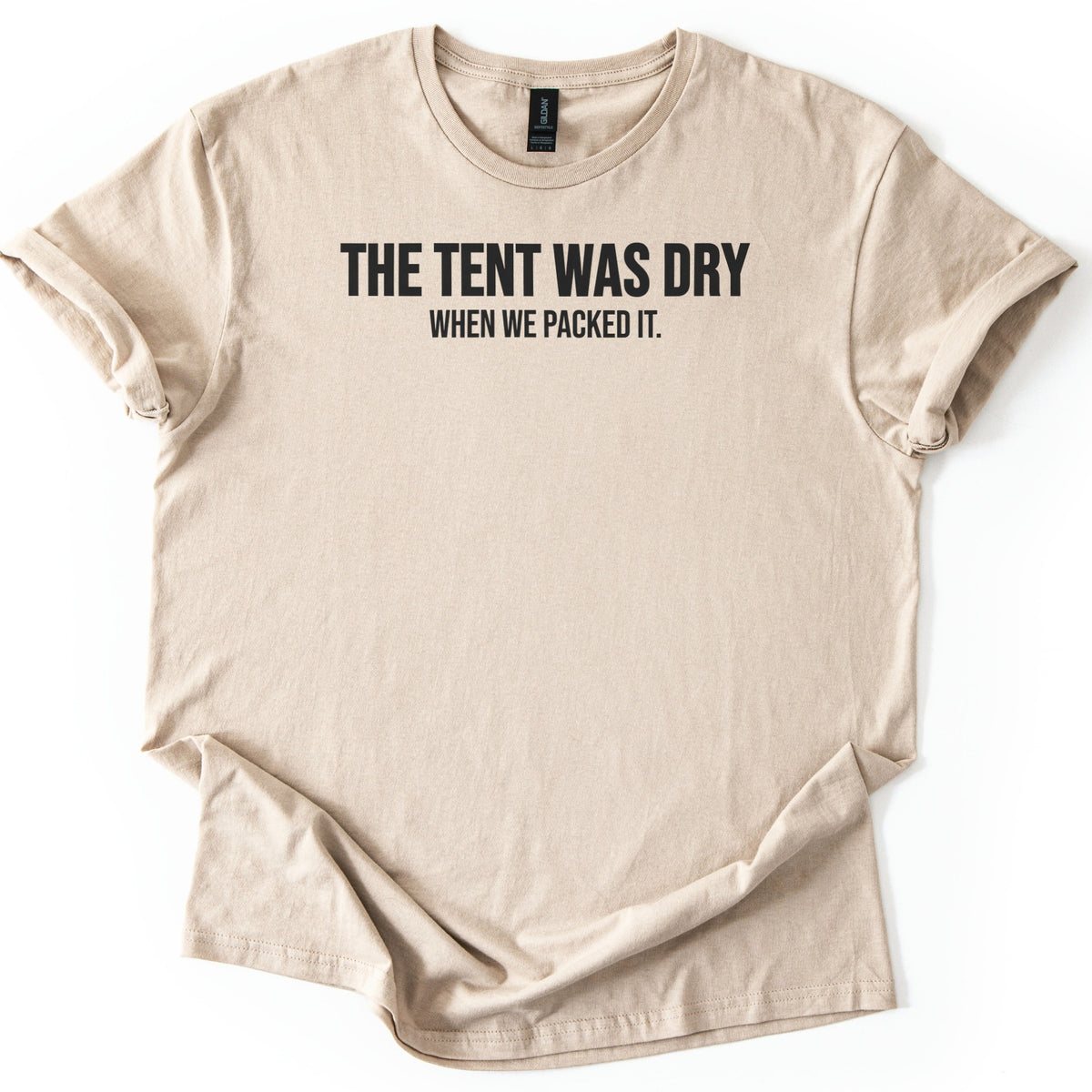 The Tent Was Dry Comfort Colors T-shirt in Sand, laid flat. Bold black capital letters across the chest read THE TENT WAS DRY, with WHEN WE PACKED IT. in smaller letters below. The shirt lies on a white surface with its sleeves rolled up.