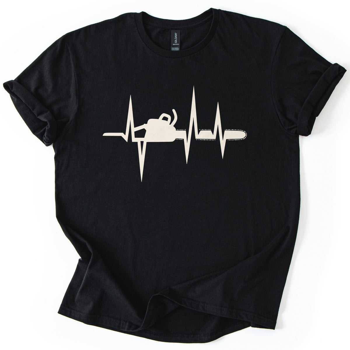 Chainsaw Heartbeat Comfort Colors T-shirt in Black, laid flat. A cream heartbeat line with sharp peaks runs across the chest, with a chainsaw silhouette set into the line, its bar and chain extending to the right. The sleeves are rolled on a white background.