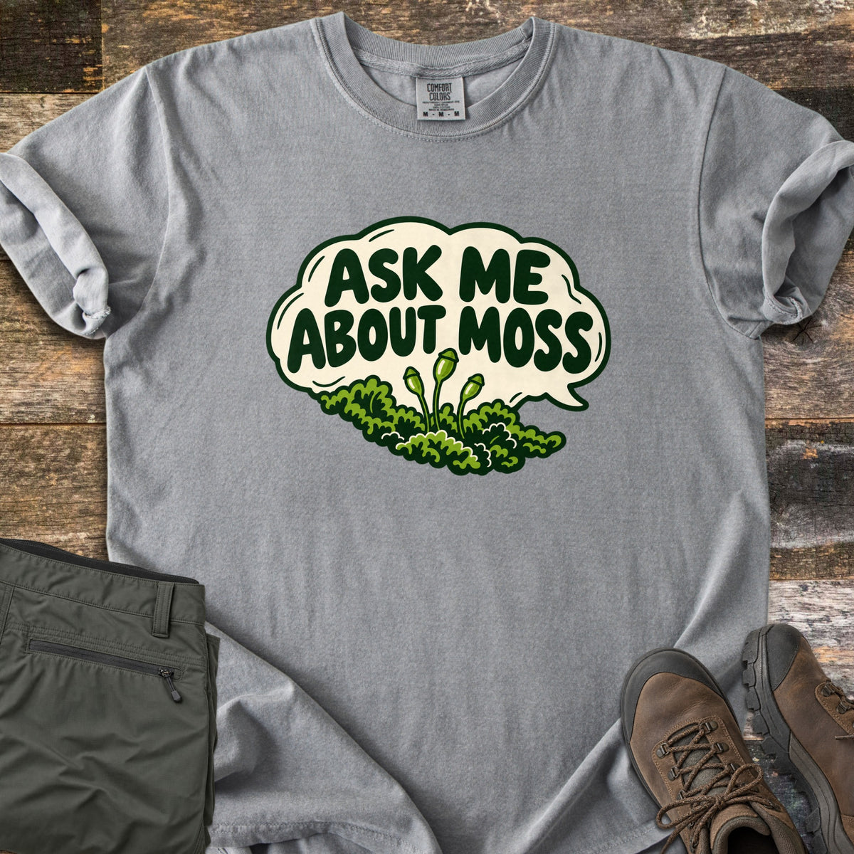 Ask Me About Moss Comfort Colors T-shirt in Grey, laid flat. A cream speech bubble reads Ask Me About Moss in dark green letters above a mound of green moss with three spore stalks. Sleeves rolled, on weathered wood with folded olive hiking pants and brown hiking boots.