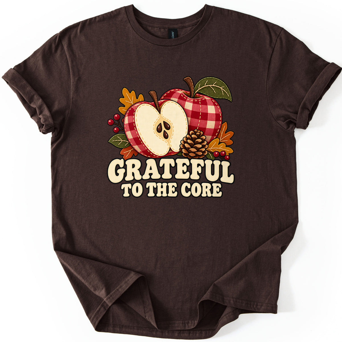 Grateful to the Core Comfort Colors T-shirt in Dark Chocolate, laid flat. Two gingham apples, one halved with brown seeds, sit among a pinecone, orange leaves and red berries over cream retro letters reading Grateful to the Core. Sleeves cuffed, on a plain white background.