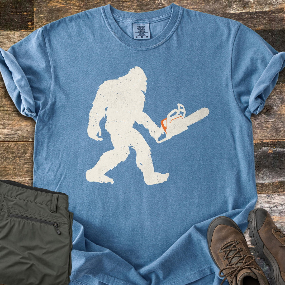 Lumber Squatch Comfort Colors T-shirt in Blue Jean, laid flat. A distressed cream silhouette of Bigfoot walks while carrying a chainsaw with orange accents. The sleeves are rolled, with folded olive pants and brown hiking boots on rustic wood planks.
