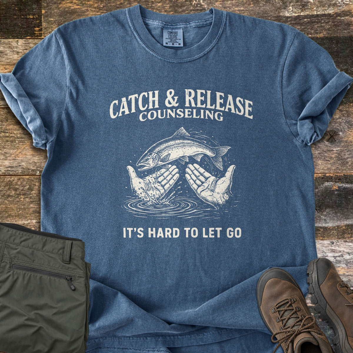 Catch & Release Counseling Comfort Colors T-shirt in Denim, laid flat. A salmon leaps between two open hands over rippling water, under the words Catch & Release Counseling and above It's Hard to Let Go. Olive pants and brown hiking boots sit beside it on weathered wood planks.