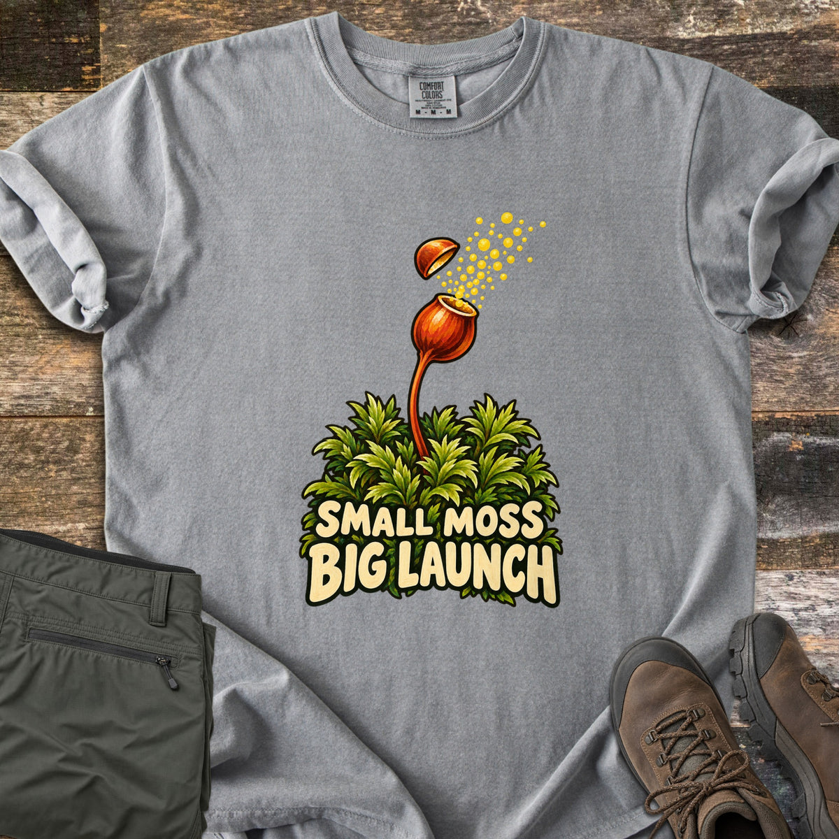 Small Moss Big Launch Comfort Colors T-shirt in Grey, laid flat. An orange moss capsule on a tall stalk launches its lid and a spray of yellow spores above a green moss clump and the words Small Moss Big Launch. Olive pants and brown hiking boots sit on weathered wood.